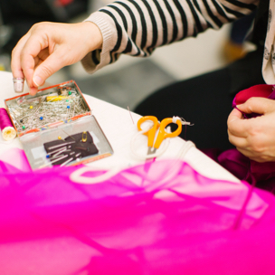 A table with pins, scissors, a pink spool of thread, and pink fabric and a person's hands pinning fabric together